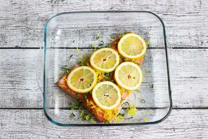 A glass baking dish contains a piece of salmon fillet topped with lemon slices and herbs.