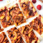 Sliced peach puff pastry tart with a flaky crust, drizzled with raspberry sauce, surrounded by fresh raspberries on parchment paper.
