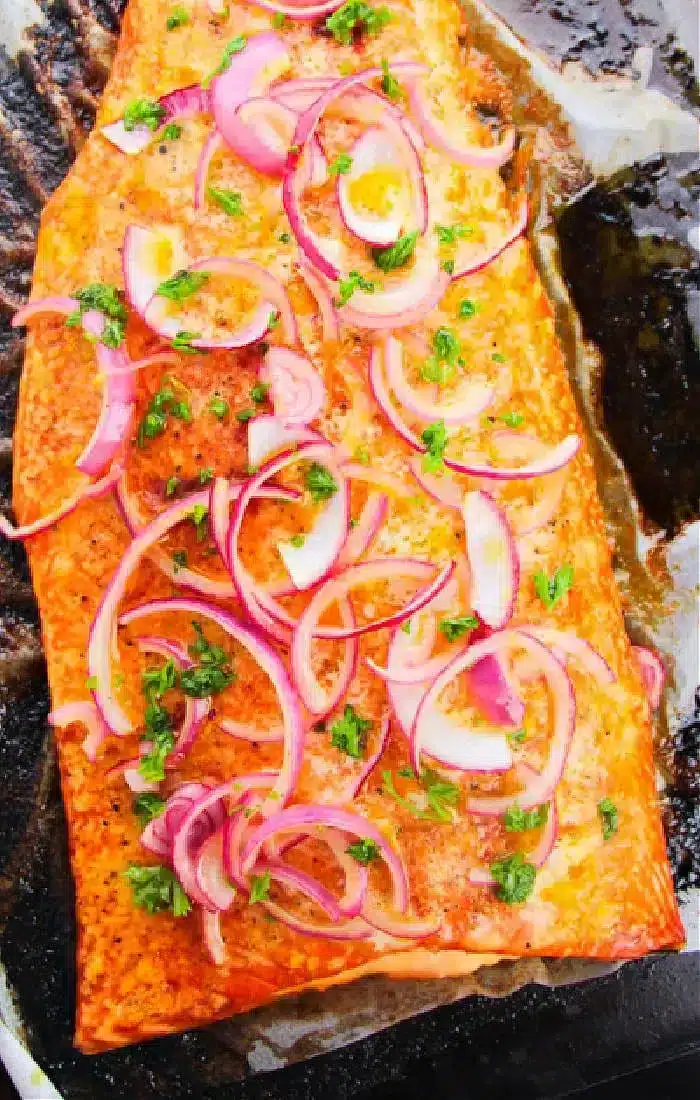 A cooked fillet of salmon on a baking tray, topped with sliced red onions and garnished with chopped green herbs.