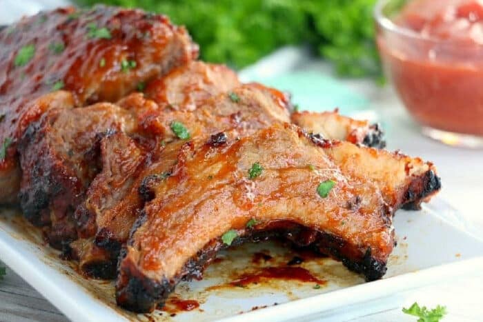 Barbecue ribs in air fryer Ninja with glaze and herbs on a white plate, served with sauce and fresh greens in the background.
