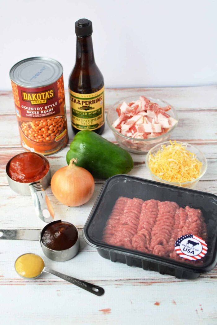 A variety of ingredients for a baked bean casserole are arranged on a white wooden surface, including a can of country-style baked beans, Worcestershire sauce, chopped bacon, a green bell pepper, shredded cheese, a ground meat package with an American flag sticker, an onion, measuring cups with sauce, and mustard.