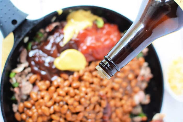 A close-up of a skillet filled with baked bean casserole, ground meat, yellow mustard, and ketchup. A bottle is seen from the top right corner, pouring additional sauce onto the mixture. The skillet handle is visible on the left side of the image.