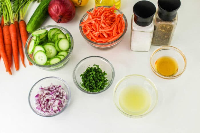 An image of assorted Carrot and Cucumber Salad ingredients and seasonings on a white surface. Items include chopped cucumbers, julienned carrots, chopped red onions, parsley, lemon juice, olive oil, and containers of salt and pepper. Additional whole vegetables are nearby.