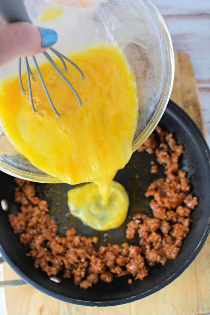 A hand is seen pouring whisked eggs from a bowl into a frying pan that contains browned ground meat, possibly chorizo. The pan rests on a wooden surface. The person is holding a whisk in the bowl, which is slightly tilted. This demonstrates how to cook chorizo and eggs effortlessly.