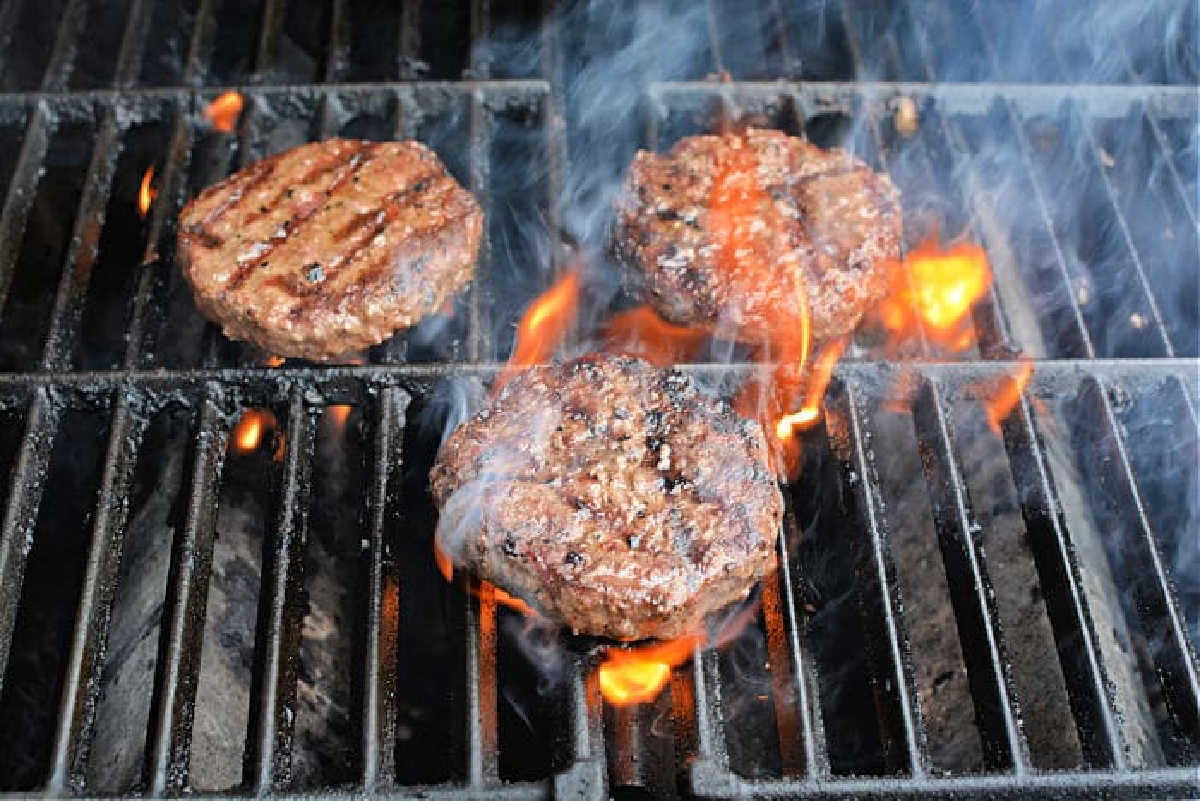 Three beef patties grilling over open flames and smoke on a barbecue grill&mdash;discover how to grill frozen burgers to perfection with this mouthwatering scene.