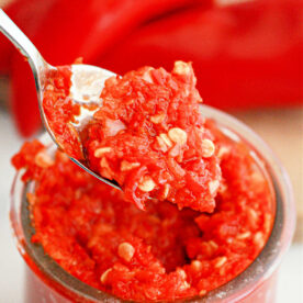 A close-up of a spoon holding a portion of red pepper paste with visible seeds, above a glass jar filled with the same rich mixture. In the background, whole red peppers are slightly out of focus, reminiscent of a savory Garlic Chili Paste recipe.