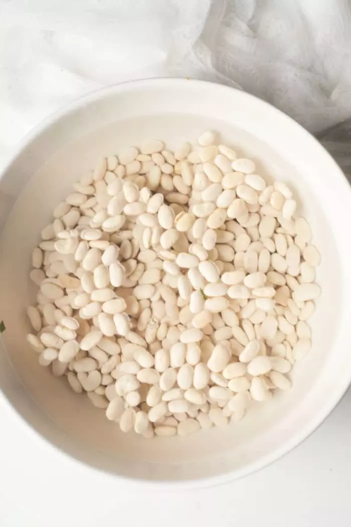 A white bowl filled with Great Northern beans and water, is placed on a light-colored surface. 