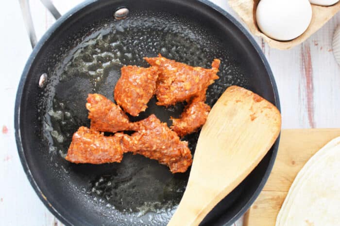 A black skillet sizzles with pieces of chorizo being cooked, a wooden spatula resting in the pan. Nearby on the counter, a carton of eggs awaits its turn, next to a tortilla on a wooden cutting board—perfect for learning how to cook chorizo and eggs.