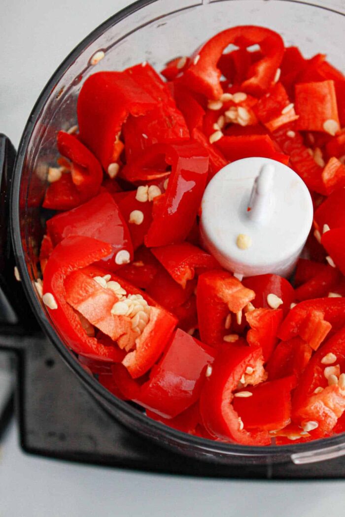 A food processor filled with cut red bell peppers, seeds, and a hint of garlic, ready for blending into a delicious chili paste recipe.