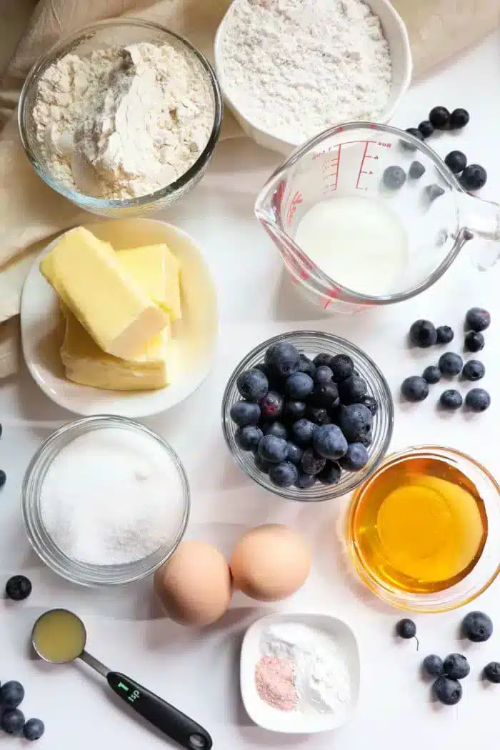 Various baking ingredients arranged on a surface include flour in a bowl, blueberry muffin tops in a dish, butter on a plate, sugar in a bowl, a measuring cup of milk, two eggs, honey in a small bowl, baking powder, and lemon juice. Blueberries are scattered around.