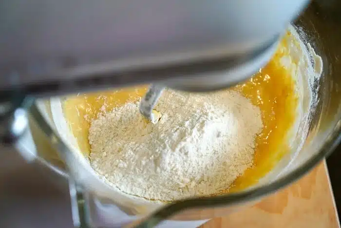 A close-up view of a stand mixer with a dough hook attachment, mixing flour and yellow batter in a metal bowl. Some dry flour is still visible on top, hinting at the transformation into delicious blueberry muffin tops.