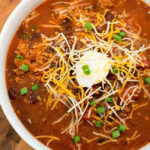 Bowl of ninja foodi chili topped with shredded cheese, sour cream, and chopped green onions on a wooden surface—a perfect mild chili recipe for cozy nights.