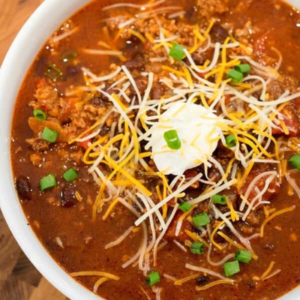 Bowl of ninja foodi chili topped with shredded cheese, sour cream, and chopped green onions on a wooden surface—a perfect mild chili recipe for cozy nights.