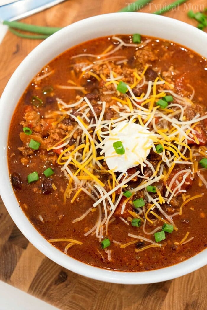 Mild chili recipe using ingredients in your pantry and fridge right now! Instructions for your Crockpot, stovetop, or pressure cooker so dinner is perfect. #chili #mild #crockpot #crockpotrecipes #instantpot #ninjafoodi #mealthy Bowl of ninja foodi chili topped with shredded cheese, sour cream, and chopped green onions on a wooden surface—a perfect mild chili recipe for cozy nights.