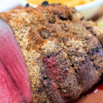 Close-up of a sliced, seasoned roast beef with a crispy crust, enhanced with Pot Roast Seasoning, with a side dish blurred in the background.