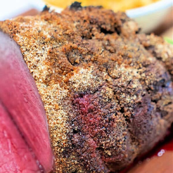Close-up of a sliced, seasoned roast beef with a crispy crust, enhanced with Pot Roast Seasoning, with a side dish blurred in the background.