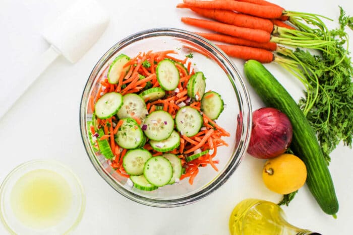 A glass bowl filled with a Carrot and Cucumber Salad, featuring sliced cucumbers, shredded carrots, and chopped herbs. Surrounding the bowl are whole carrots with greens, a cucumber, a red onion, a lemon, a small bowl of dressing, a bottle of oil, and a white kitchen tool.