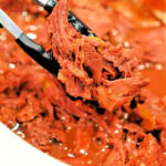 Tongs holding shredded, saucy beef chuck roast over a bowl filled with more shredded beef in sauce, perfect for the slow cooker.