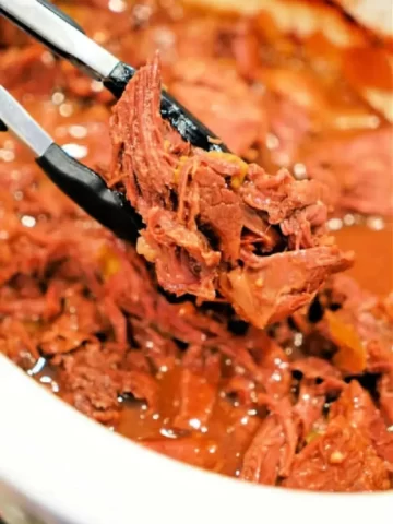 Close-up of a pair of tongs holding shredded beef chuck roast over a bowl brimming with more tender meat. The beef is cooked to perfection in a slow cooker and sits enticingly in a rich, reddish sauce.