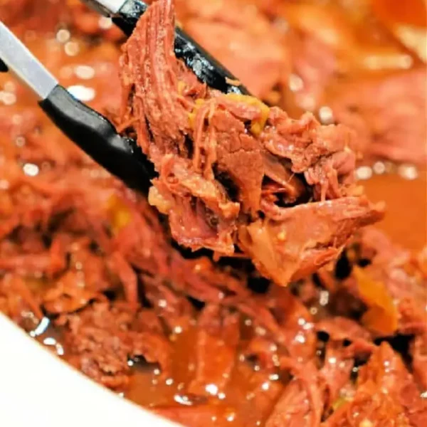 Close-up of a pair of tongs holding shredded beef chuck roast over a bowl brimming with more tender meat. The beef is cooked to perfection in a slow cooker and sits enticingly in a rich, reddish sauce.