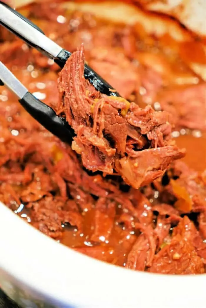 Close-up of a pair of tongs holding shredded beef chuck roast over a bowl brimming with more tender meat. The beef is cooked to perfection in a slow cooker and sits enticingly in a rich, reddish sauce.