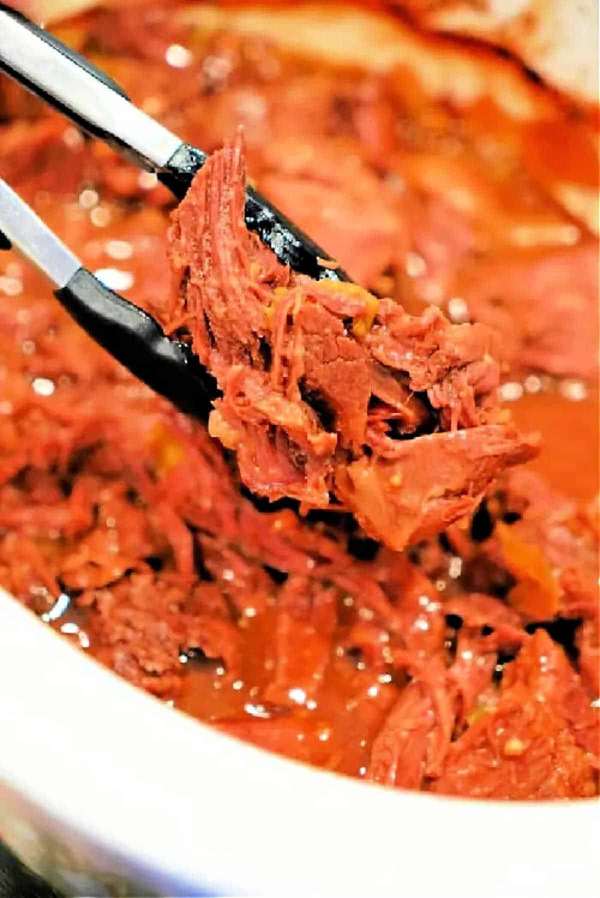Tongs holding shredded, saucy beef chuck roast over a bowl filled with more shredded beef in sauce, perfect for the slow cooker.