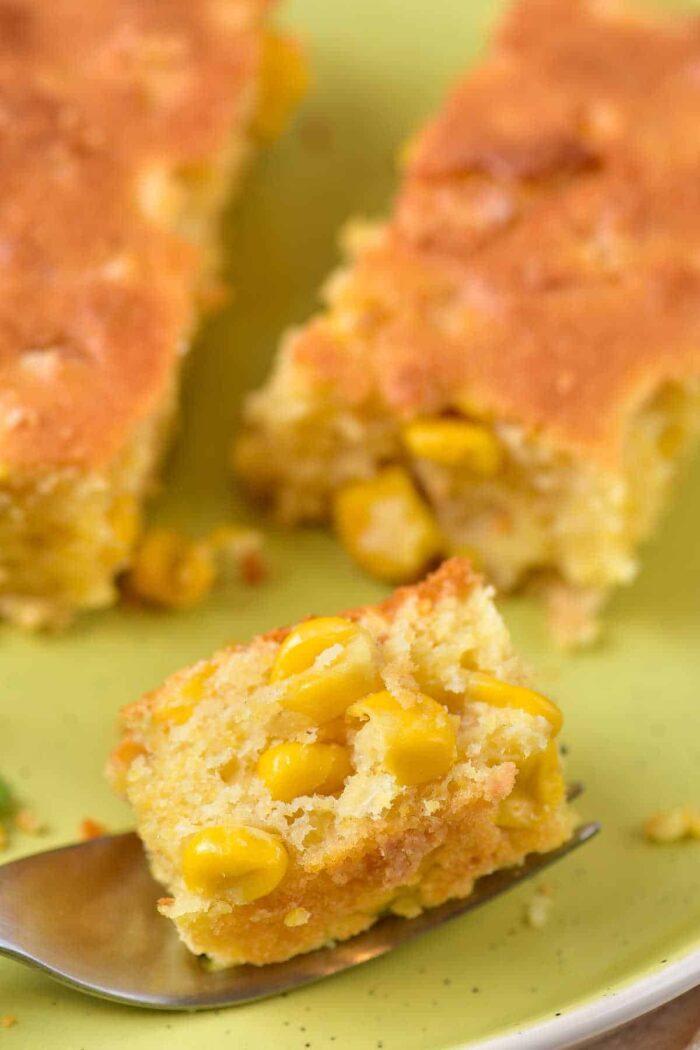 A close-up of a piece of corn casserole with corn kernels, reminiscent of corn pudding without Jiffy mix, placed on a green plate. A fork holding a smaller piece of the cornbread is visible in the foreground.