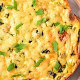 A close-up of a Cottage Cheese Quiche on a white plate. The quiche has a golden-brown crust, topped with melted cheese, small bits of vegetables, and garnished with chopped green onions.