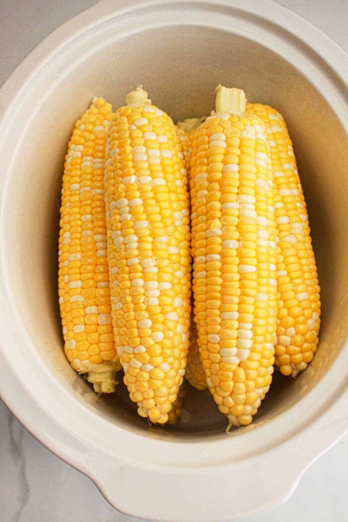 Four ears of corn are placed inside a white crockpot. The corn on the cob appears to be fresh with kernels that are a mix of yellow and white. The slow cooker is situated on a light-colored surface.