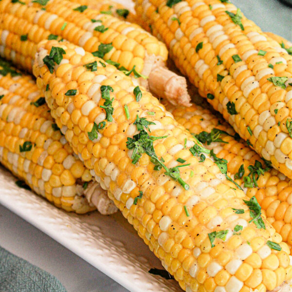 A close-up of several ears of Crockpot Corn on the Cob placed on a white rectangular dish. The corn is garnished with chopped fresh herbs and a sprinkle of seasoning. A green cloth is partially visible in the background.