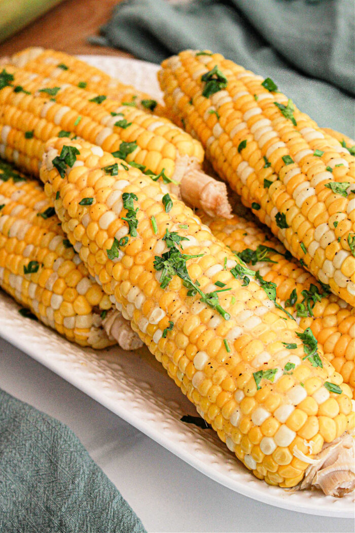 A close-up of several ears of Crockpot Corn on the Cob placed on a white rectangular dish. The corn is garnished with chopped fresh herbs and a sprinkle of seasoning. A green cloth is partially visible in the background.