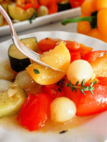 A close-up of a plate with cooked vegetables, including cherry tomatoes, onions, and slices of zucchini. A fork is picking up a piece of yellow pepper. A yellow bell pepper and more slow cooker vegetables are visible in the background.