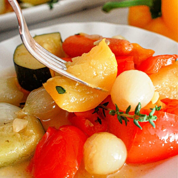 A close-up of a plate with cooked vegetables, including cherry tomatoes, onions, and slices of zucchini. A fork is picking up a piece of yellow pepper. A yellow bell pepper and more slow cooker vegetables are visible in the background.