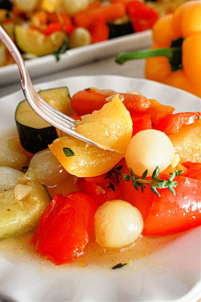 A close-up of a plate with cooked vegetables, including cherry tomatoes, onions, and slices of zucchini. A fork is picking up a piece of yellow pepper. A yellow bell pepper and more slow cooker vegetables are visible in the background.