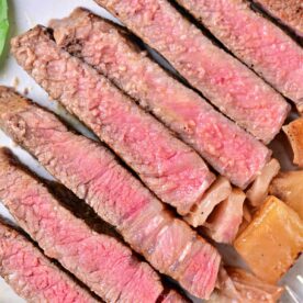 A plate with several slices of medium-rare beef strip loin steaks arranged in a line. The meat is pink in the center with a browned outer edge, featuring a juicy texture. Some greens are visible in the corner of the plate, showing you how to cook beef strip loin steaks to perfection.