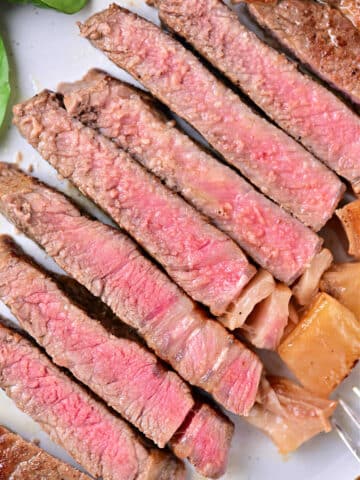 A plate with several slices of medium-rare beef strip loin steaks arranged in a line. The meat is pink in the center with a browned outer edge, featuring a juicy texture. Some greens are visible in the corner of the plate, showing you how to cook beef strip loin steaks to perfection.