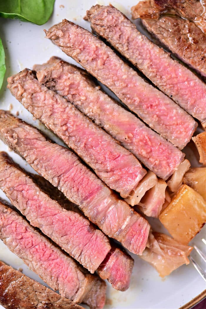 A plate with several slices of medium-rare beef strip loin steaks arranged in a line. The meat is pink in the center with a browned outer edge, featuring a juicy texture. Some greens are visible in the corner of the plate, showing you how to cook beef strip loin steaks to perfection.