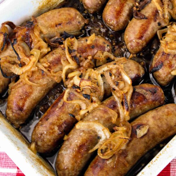 A rectangular baking dish filled with cooked sausages topped with caramelized onions sits enticingly on a red and white checkered tablecloth, making you wonder how to cook bratwurst to perfection.