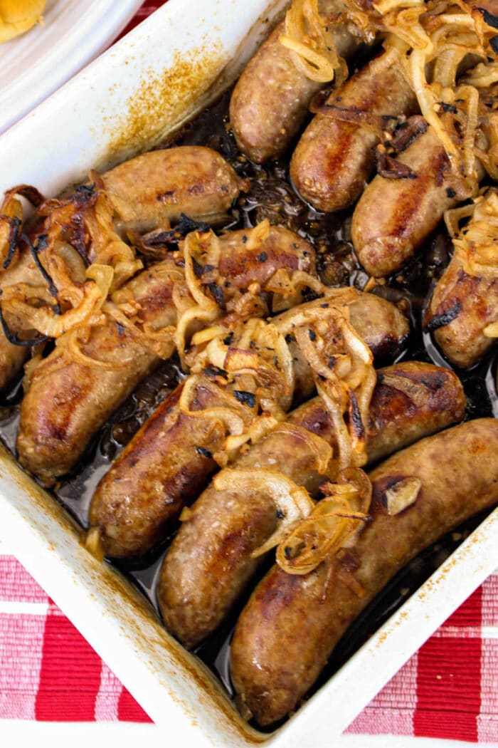 A rectangular baking dish filled with cooked sausages topped with caramelized onions sits enticingly on a red and white checkered tablecloth, making you wonder how to cook bratwurst to perfection.