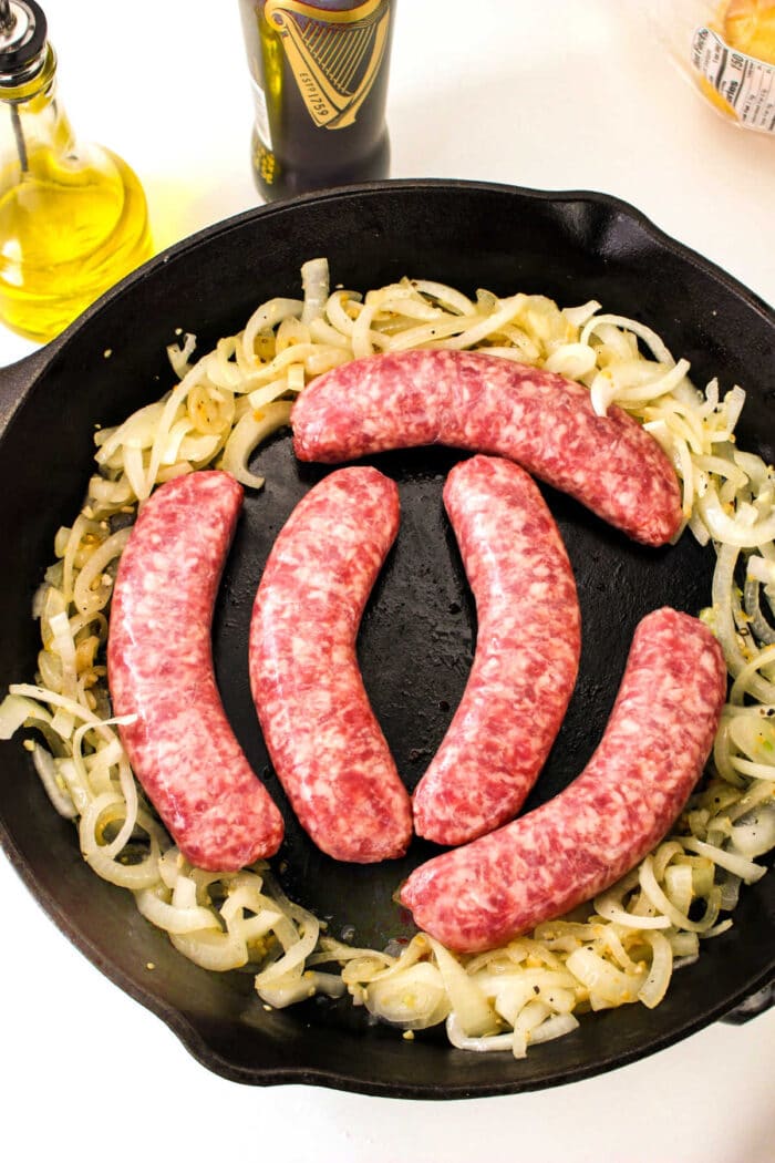 Five raw sausages are placed on a bed of sliced onions sizzling in a cast iron skillet, demonstrating how to cook bratwurst to perfection. A bottle of olive oil and a bottle of Guinness beer are visible in the background.