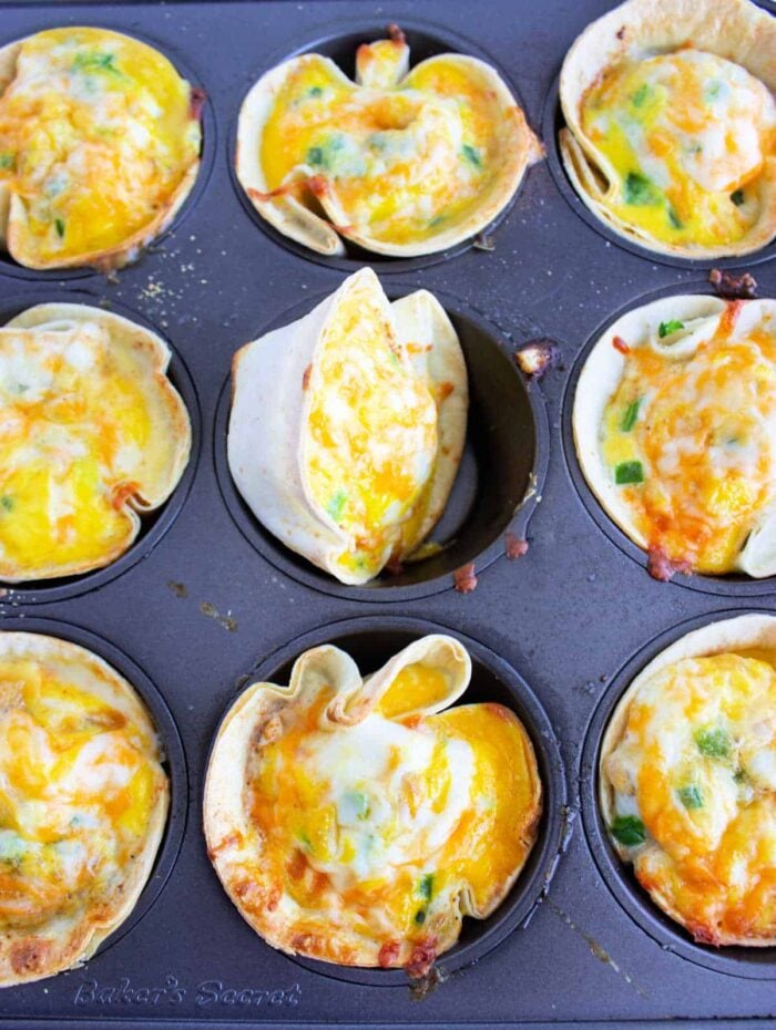 A muffin tin containing nine baked tortilla cups filled with a delightful mixture of melted cheese and diced green peppers, reminiscent of cottage cheese quiche. One of the cups appears slightly overfilled and has spilled out. The tin is marked with "Baker's Secret" at the bottom left.