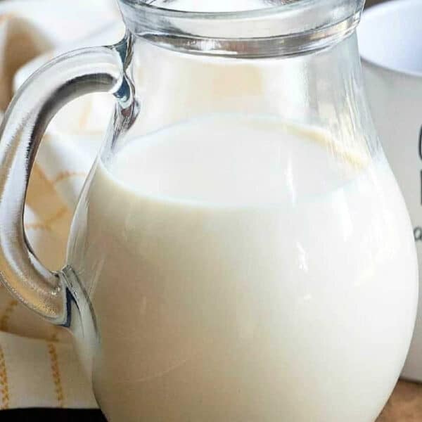 A clear glass pitcher filled with milk and pumpkin spice coffee creamer sits on a wooden table next to a white cloth.