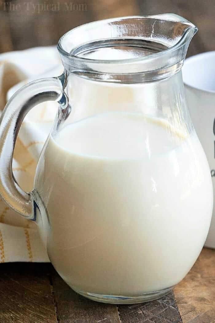 A clear glass pitcher filled with milk and pumpkin spice coffee creamer sits on a wooden table next to a white cloth.
