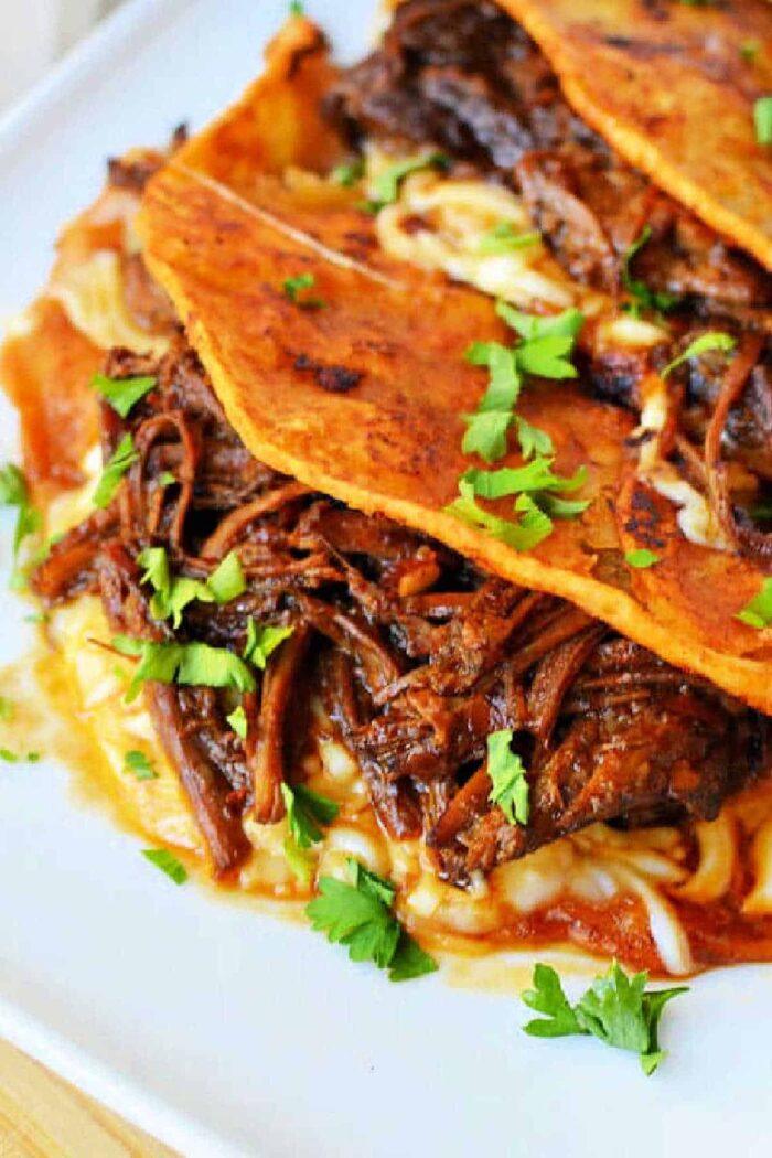 A close-up of three short rib tacos on a white plate. The tacos are filled with shredded meat and melted cheese, topped with chopped fresh cilantro. A bowl of dipping sauce is partially visible in the background.