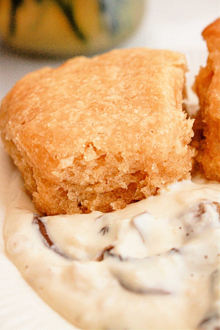 Close-up of a biscuit with a golden-brown crust placed on top of white gravy with visible bits of mushroom. The texture of the biscuit looks flaky and crumbly, while the gravy appears creamy and thick. This Slow Cooker Biscuits and Gravy dish promises comfort in every bite.