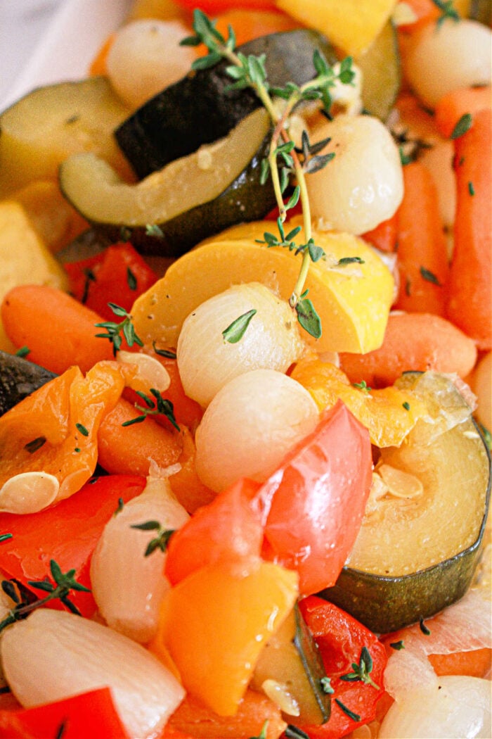 A close-up of a mixed vegetable medley featuring zucchini slices, baby carrots, pearl onions, and bell pepper pieces, garnished with sprigs of thyme. The Slow Cooker Vegetables appear to be cooked and seasoned, displaying vibrant colors.