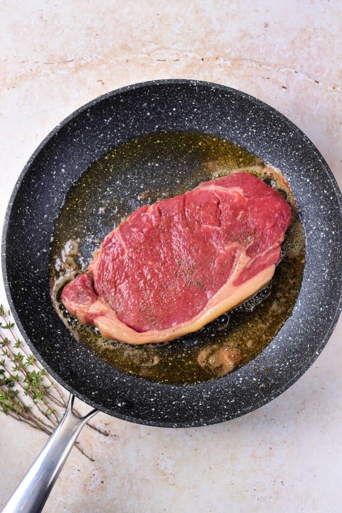 A raw steak, perfect for those learning how to cook beef strip loin steaks, is shown lying in a frying pan with some oil. The surrounding surface is a light, textured material, and a small bunch of herbs, possibly thyme, lies next to the pan. The steak appears to be in the early stages of cooking.