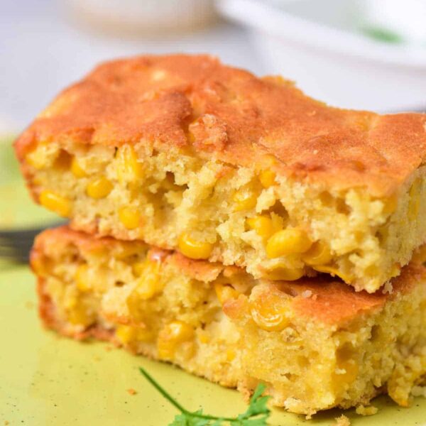 Two pieces of cornbread with visible corn kernels stacked on a light green plate remind one of traditional Corn Pudding Without Jiffy Mix. A piece of parsley adorns the plate, and a fork is partially visible in the background.