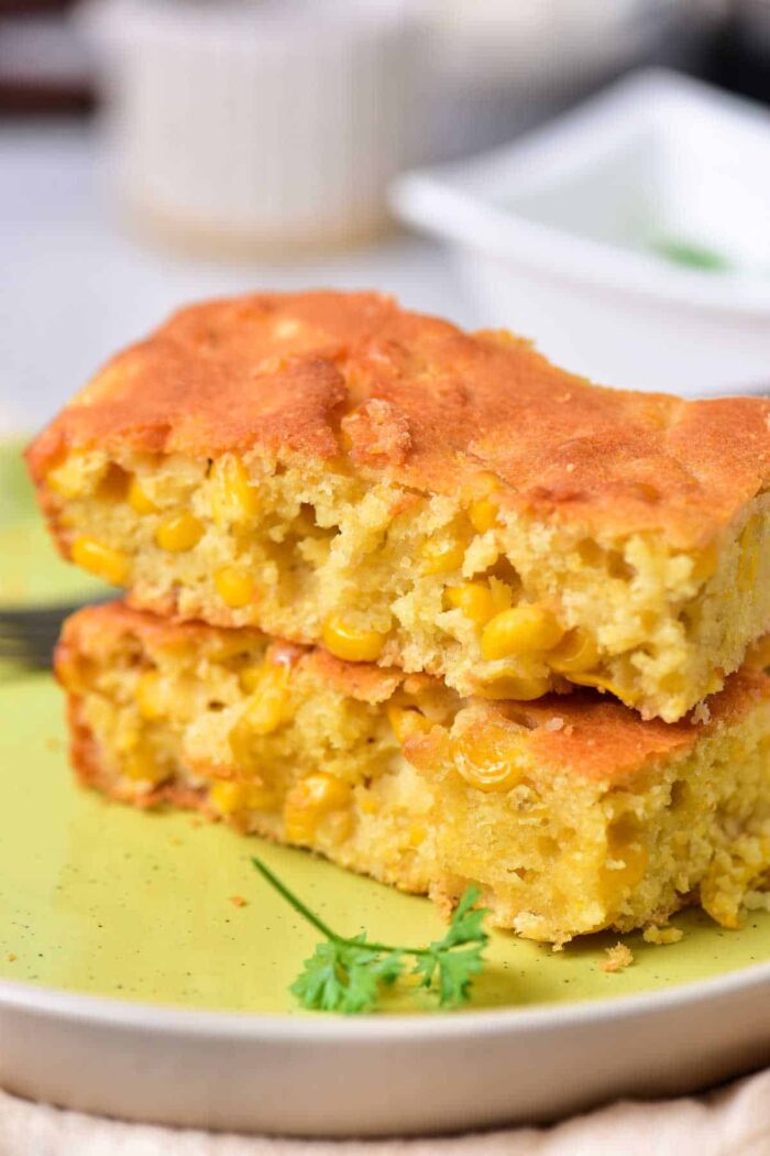 Two pieces of cornbread with visible corn kernels stacked on a light green plate remind one of traditional Corn Pudding Without Jiffy Mix. A piece of parsley adorns the plate, and a fork is partially visible in the background.