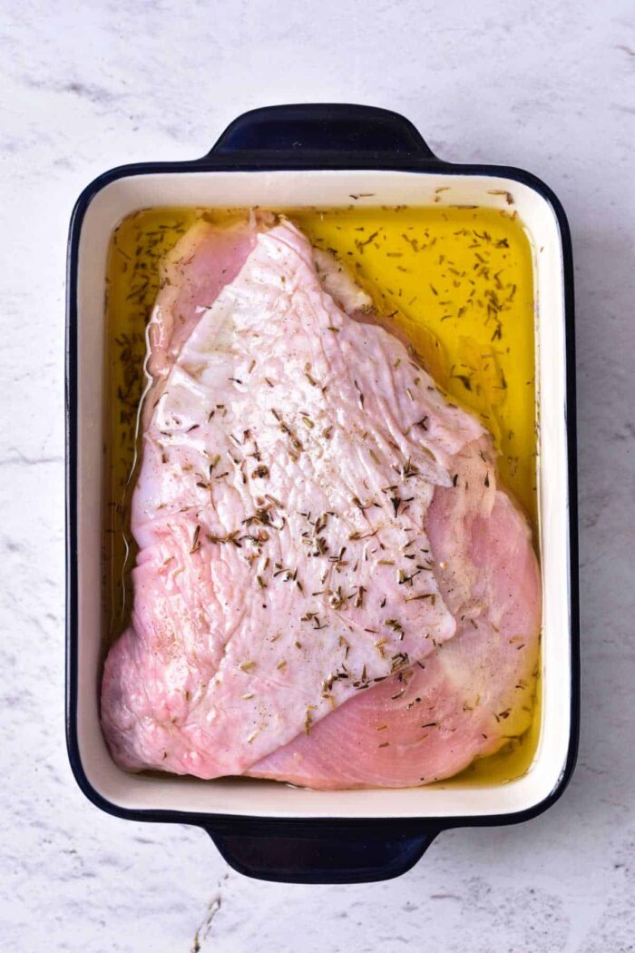 A raw turkey breast marinating in oil and seasonings, destined for a delicious turkey confit, sits in a rectangular white and black roasting pan on a light-colored surface.